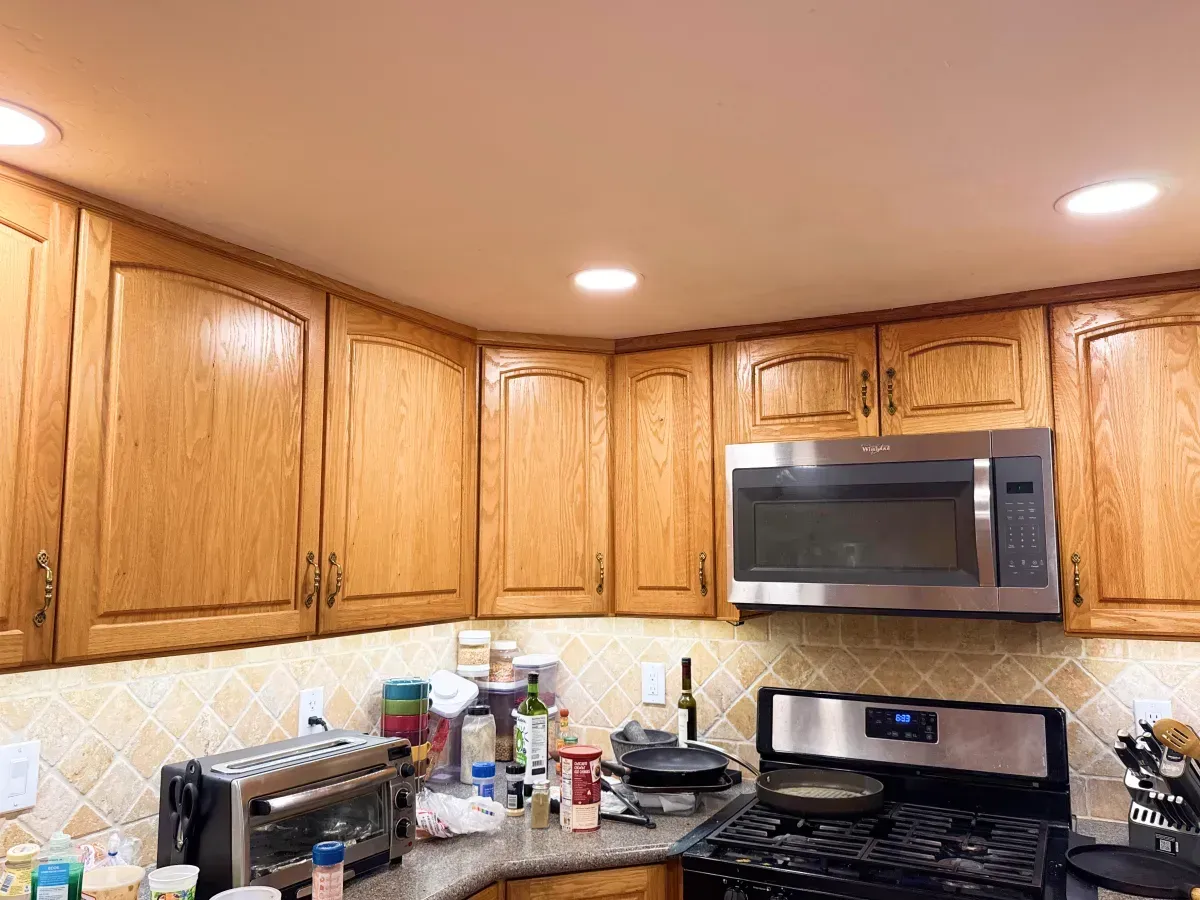 Kitchen with wooden cabinets, microwave, and stovetop. Beige backsplash and recessed ceiling lights.