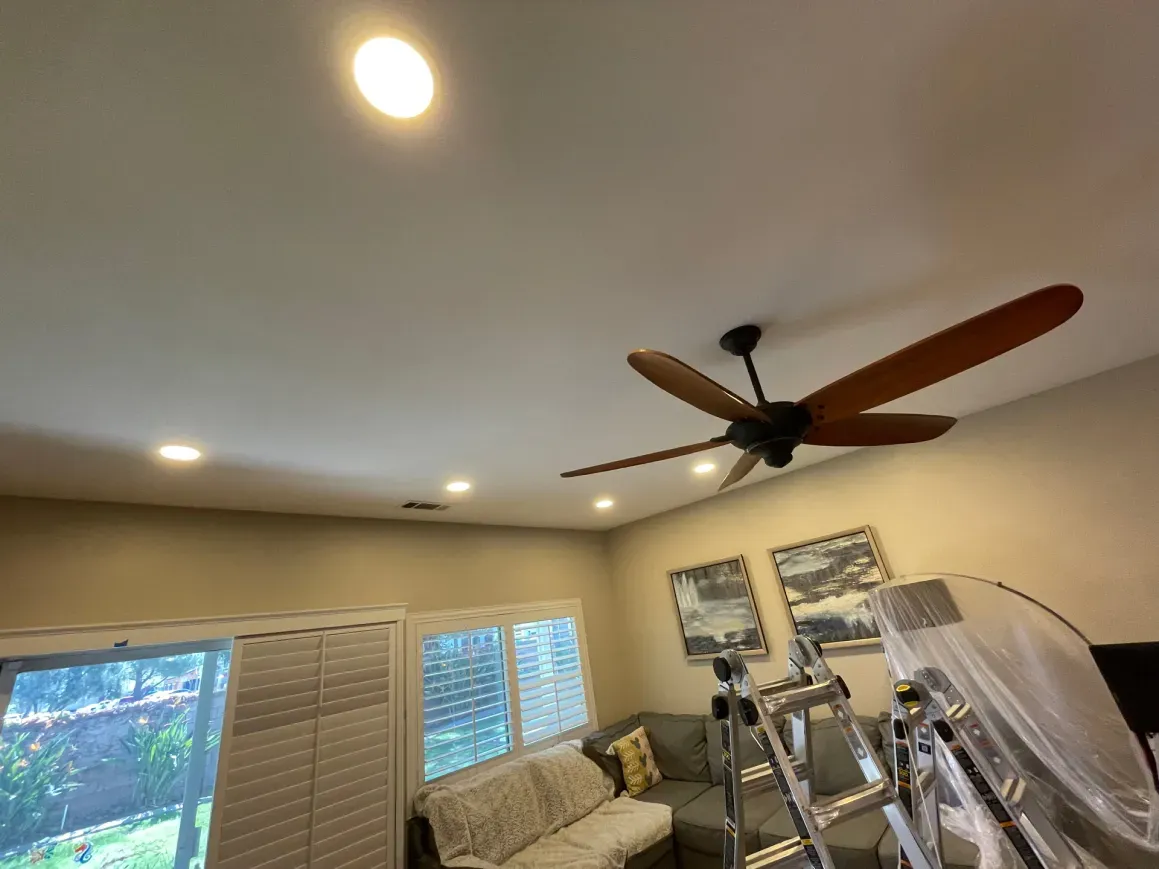 Ceiling with fan and recessed lights in a room with windows and a ladder.