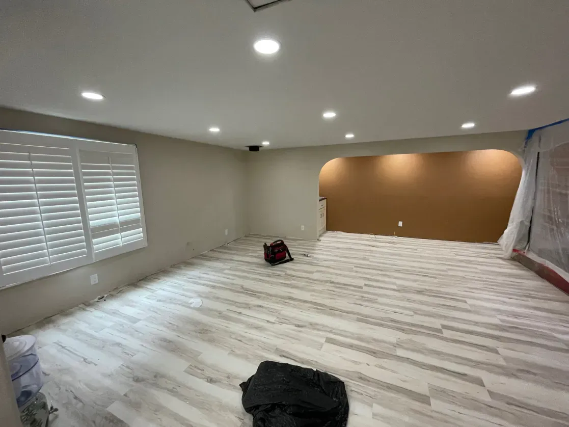 Spacious room with new wood-look flooring, recessed lighting, and a painted tan accent wall. Window with blinds on left.