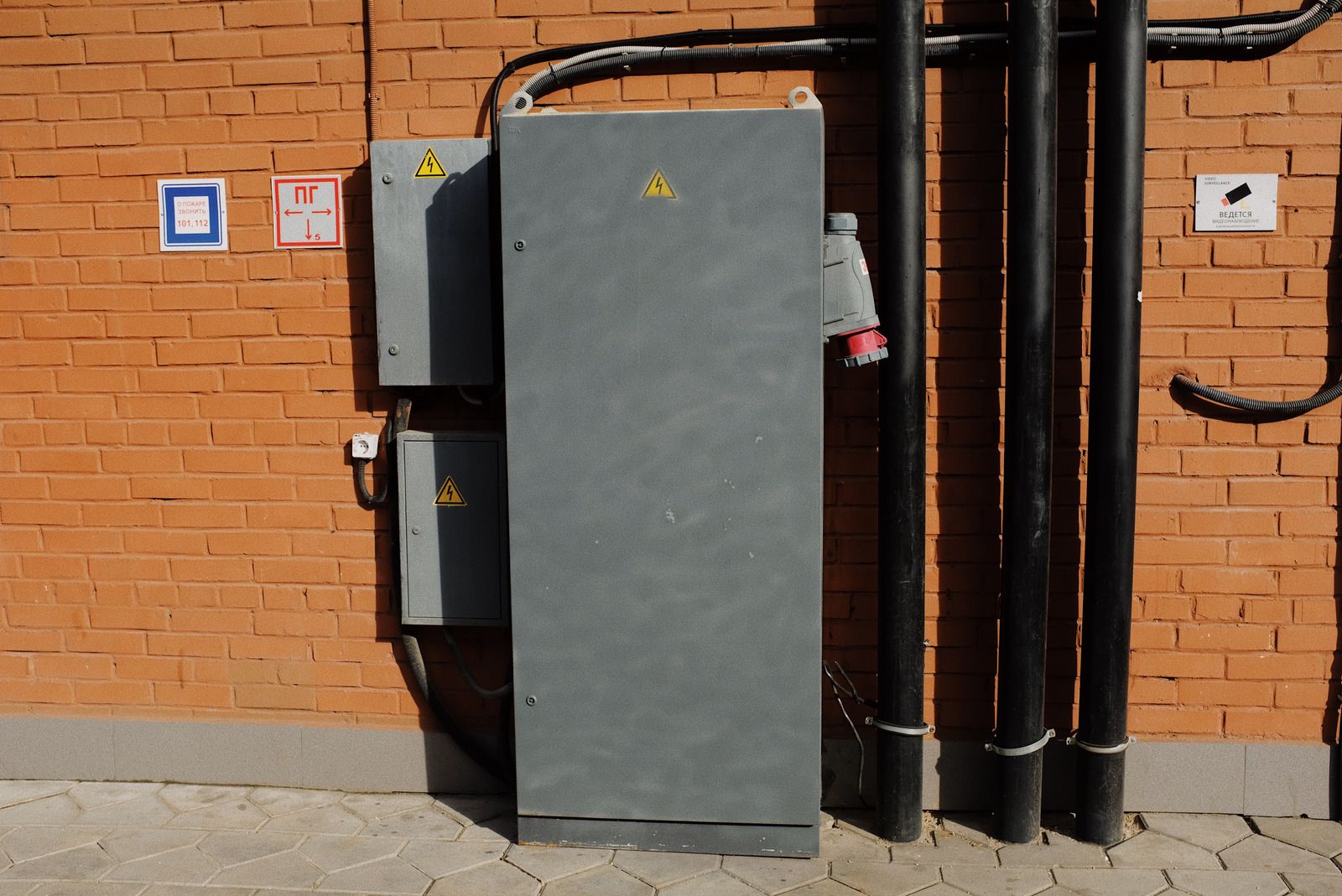 Gray electrical box on brick wall with conduit pipes, warning symbols, and a power outlet.