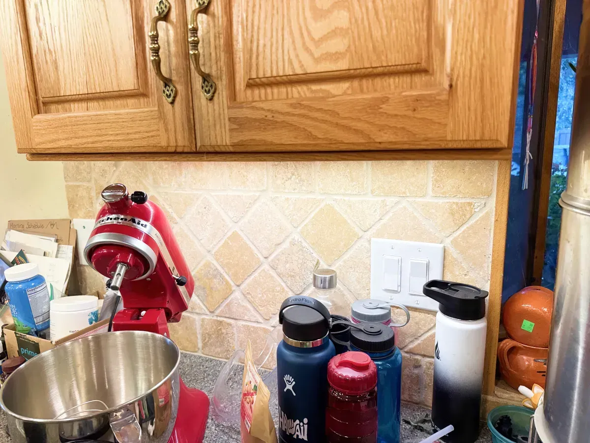 Kitchen countertop with a red stand mixer, water bottles, and a light switch beneath wooden cabinets.