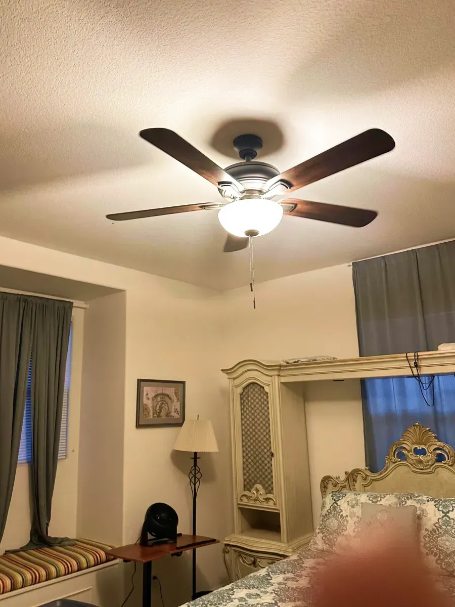 Ceiling fan with light in a bedroom. Brown blades, white ceiling. Bed, cabinet, and curtains are also visible.