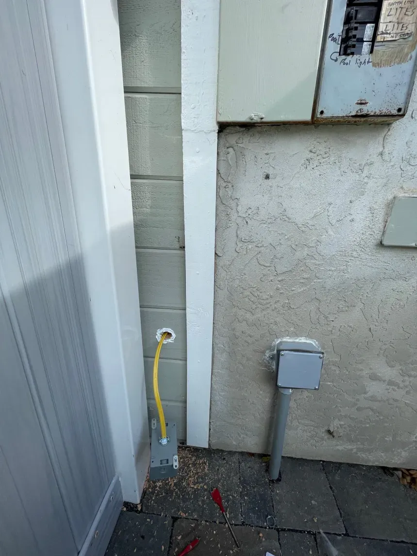 Yellow electrical wire extending from a grey electrical box on a concrete block wall, next to a white fence.