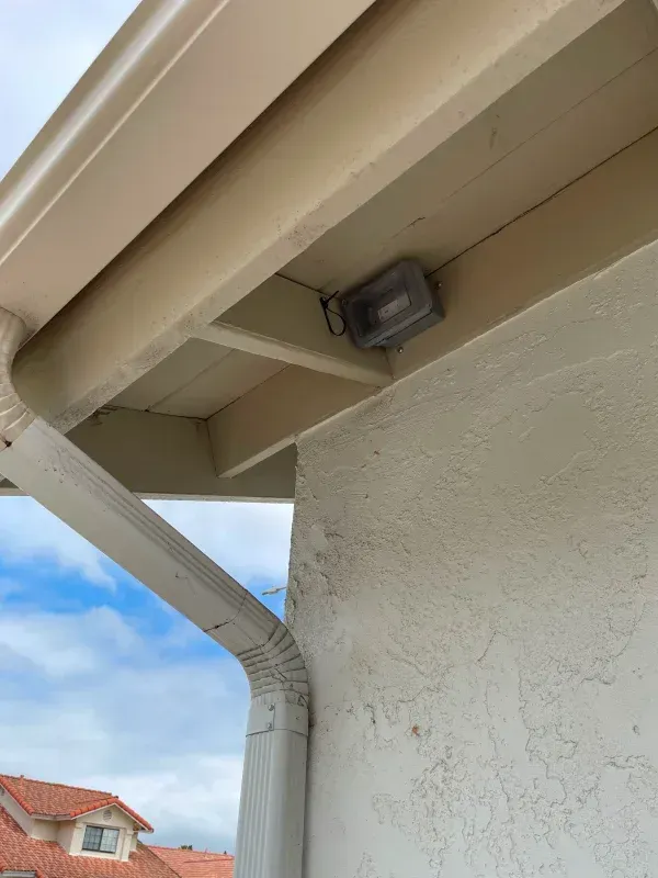 Outdoor floodlight mounted on a house's soffit, near a gutter and stucco wall.