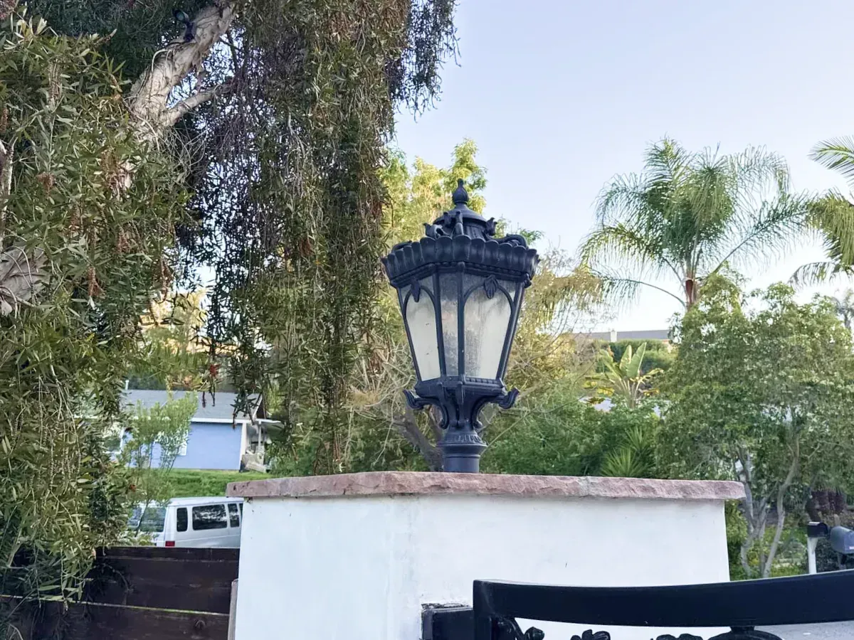 Black ornate lamp atop a white wall, surrounded by trees and a glimpse of a house.