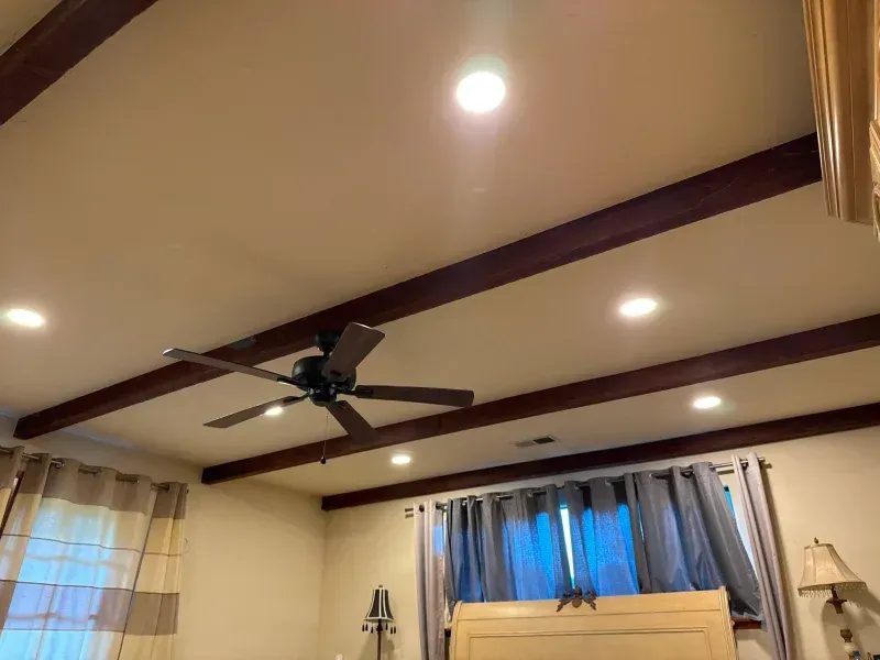 Beige ceiling with dark brown beams, recessed lights, and a ceiling fan over a bed.