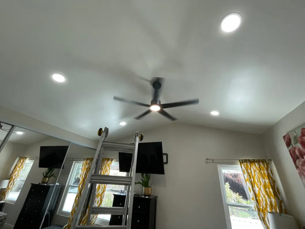 Ceiling fan installation; a ladder beneath the fan. Recessed lights and a floral curtain are also visible.