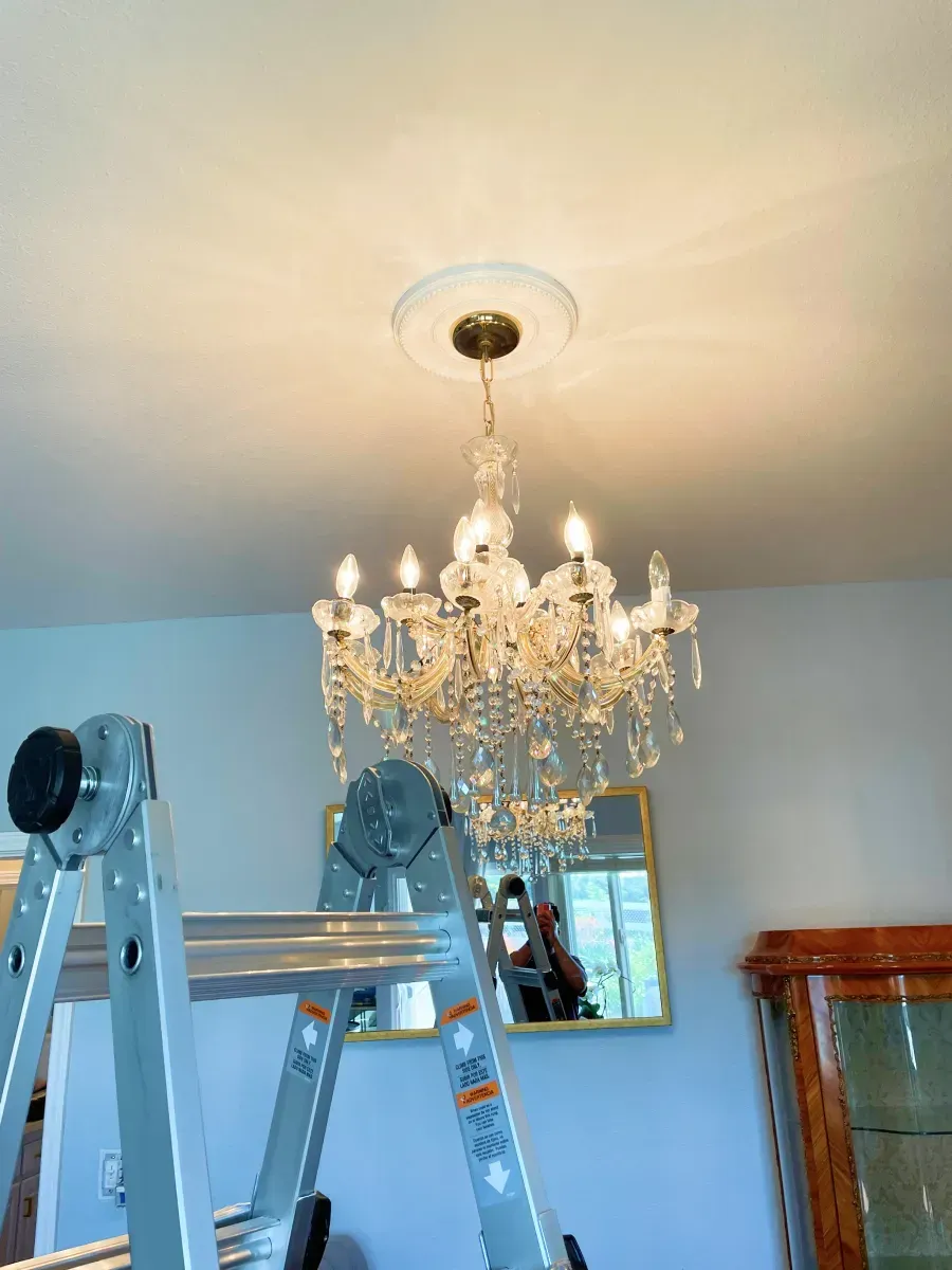 Crystal chandelier hanging from a white ceiling with a person on a ladder.