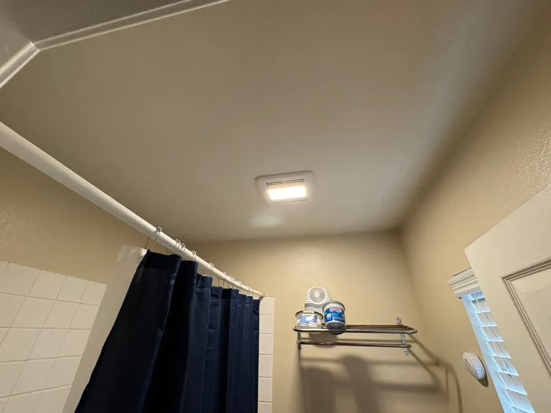 Bathroom interior: shower curtain, overhead light, towel rack with supplies, beige walls.