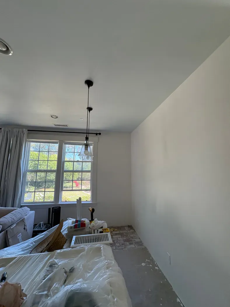 Interior view of a room with white walls, a window, and a hanging light fixture. Construction materials are visible.