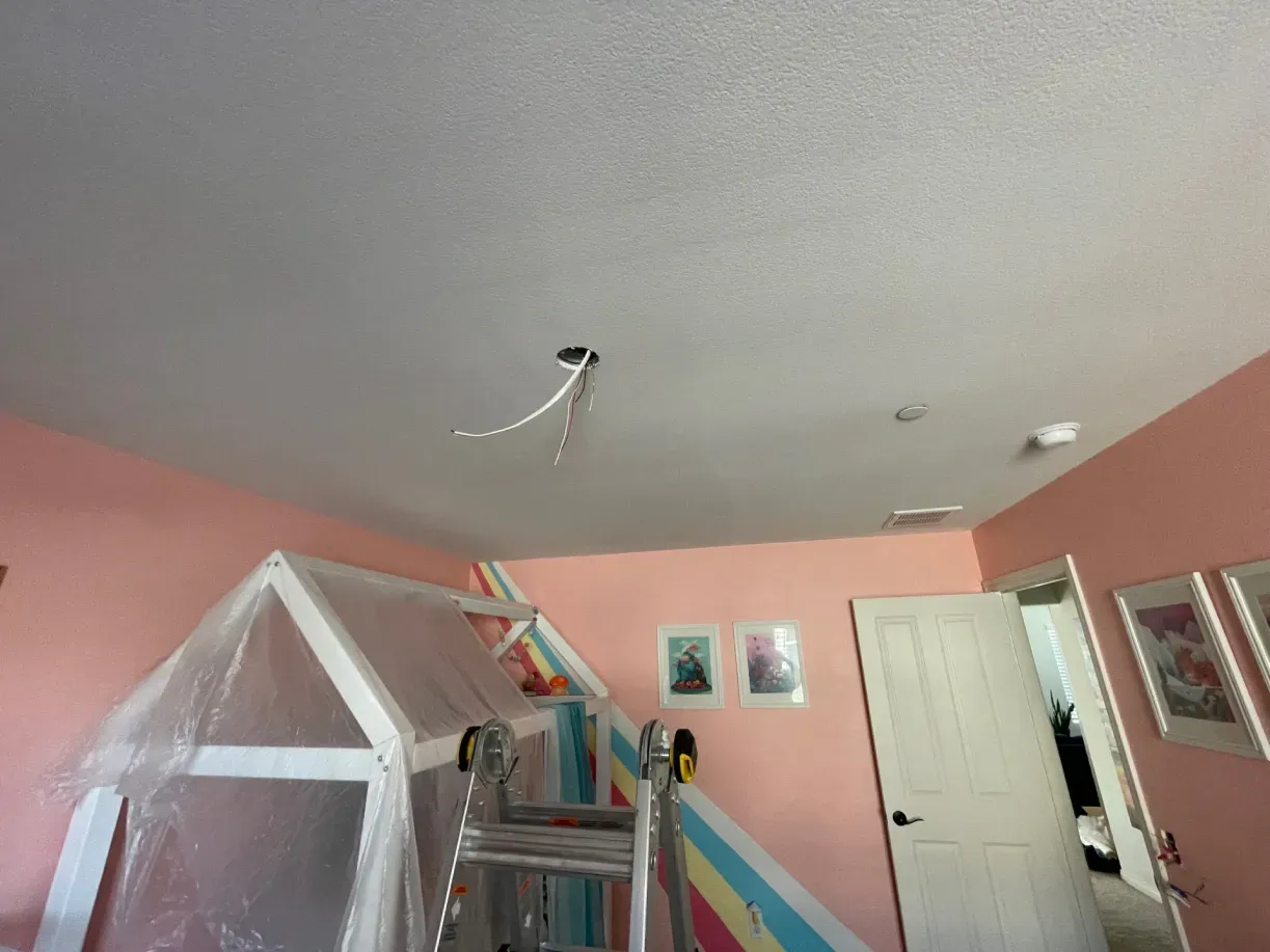 Pink-walled room with a playhouse and ladder, a light fixture and wires extend from the ceiling.