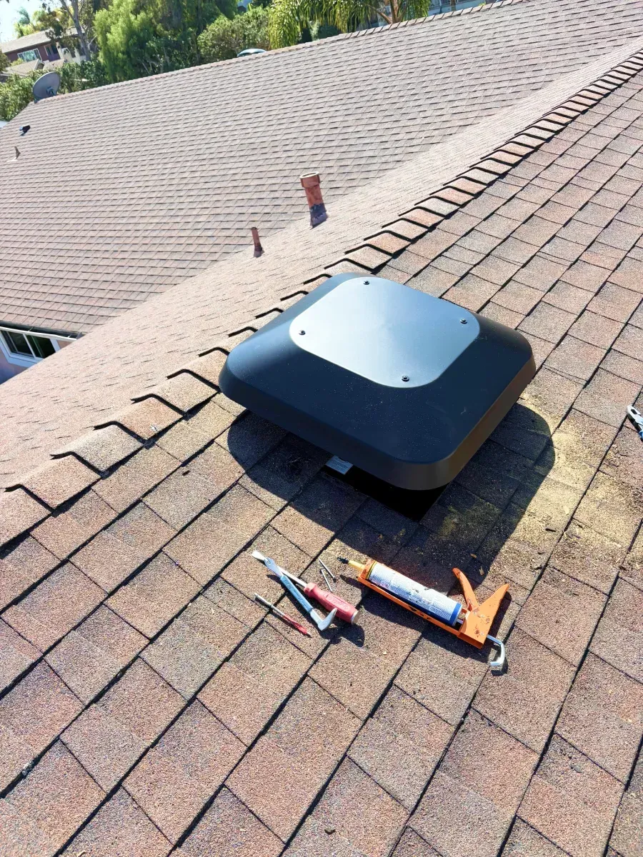 Black roof vent on a brown shingle roof, with a caulk gun and tools, outdoors.