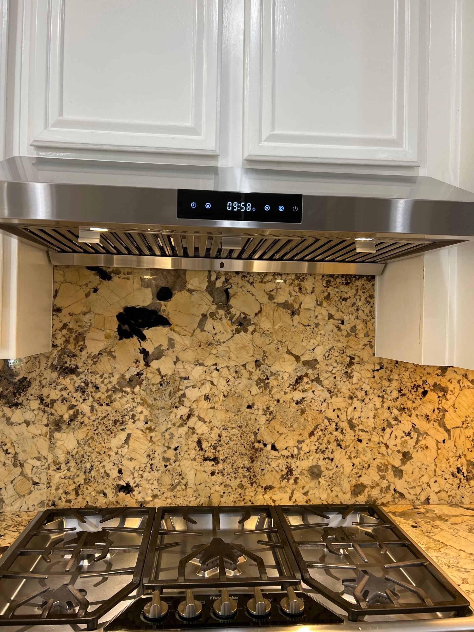 Stainless steel range hood above a gas stovetop and granite backsplash, white cabinets overhead.