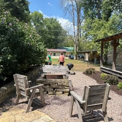 Two people grilling by a fire pit in a backyard with wooden chairs and landscaping.