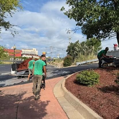 Landscapers in green shirts spread mulch along a sidewalk next to a road.