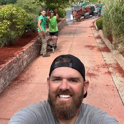 Man taking selfie, landscaping crew in background spreading mulch on pathway.