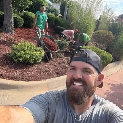 Man smiling, taking selfie with landscaping crew mulching flowerbeds.