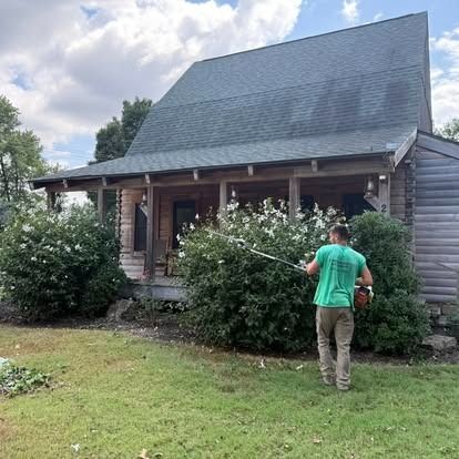 Man trims bushes in front of a wooden house. Green shirt and pants, yard with green grass.