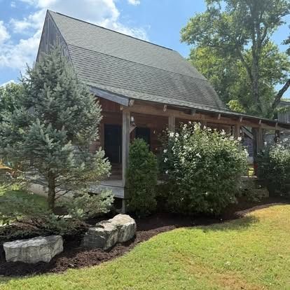 A rustic wooden cabin with a porch, surrounded by greenery and landscaping, under a sunny sky.