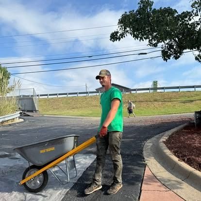 Man pushing a wheelbarrow on a paved area. He wears a green shirt, hat, and work pants. Blue sky background.