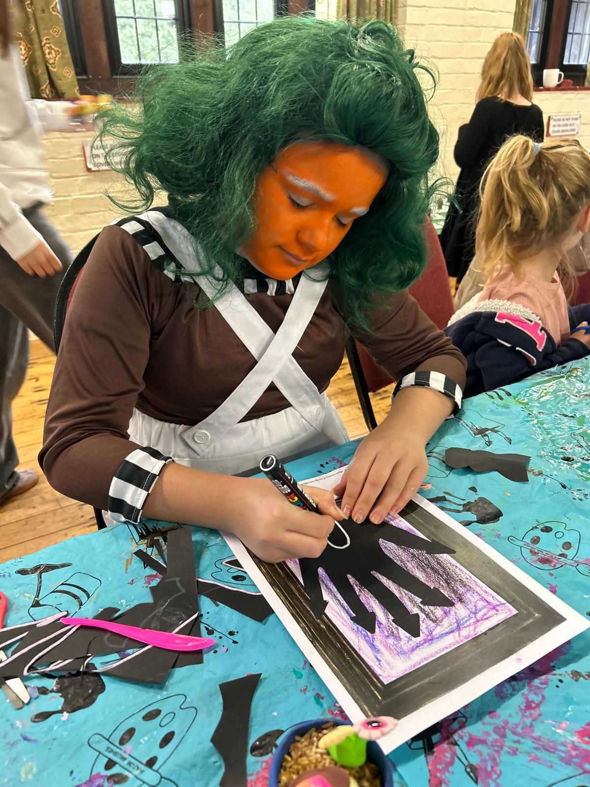 A girl with green hair and orange face paint is sitting at a table at Dragonfly crafts party