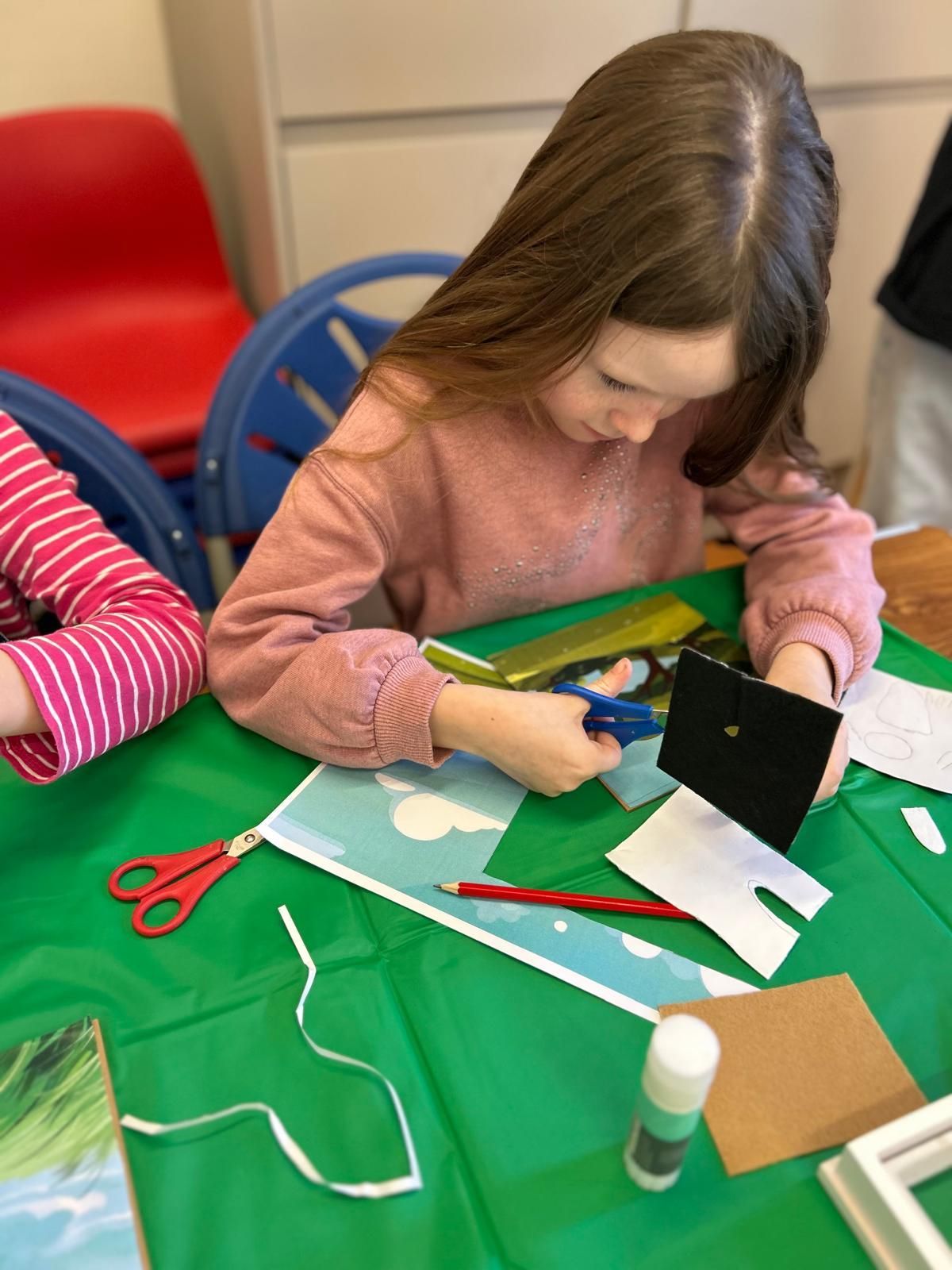 A girl is sitting at a table cutting a piece of paper with scissors at Dragonfly-craft parties.
