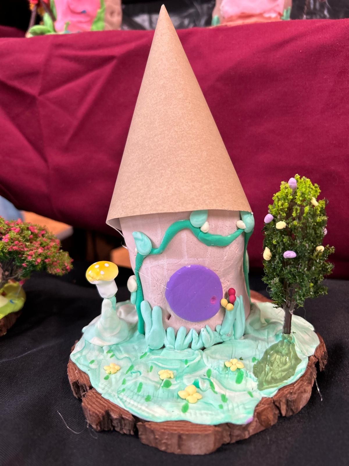 A gingerbread house with a purple door is on a wooden stump for Dragonfly crafts party