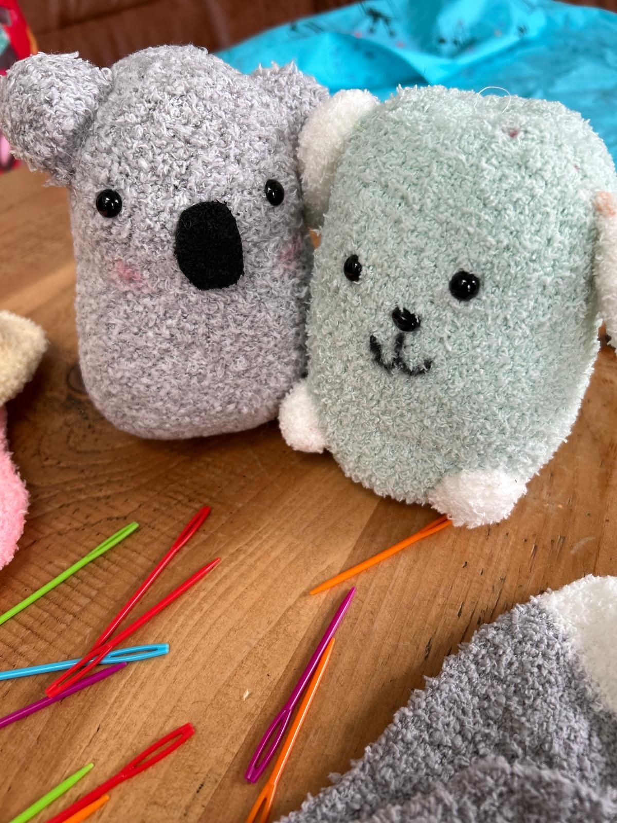 Two stuffed koalas are sitting on a wooden table next to knitting needles, for Dragonfly-craft parties