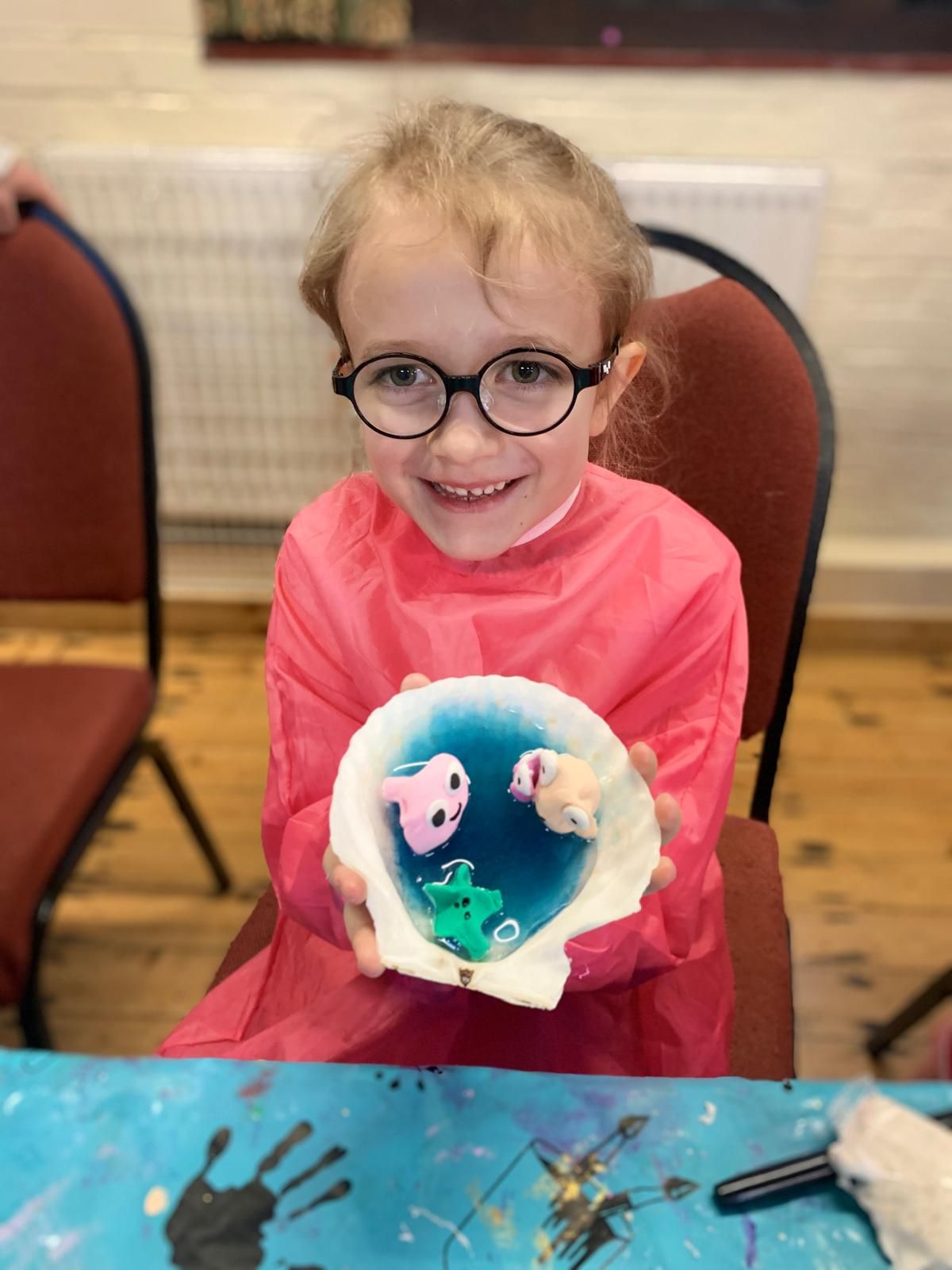 A little girl wearing glasses is holding a shell with fish in it, at Dragonfly-craft parties.