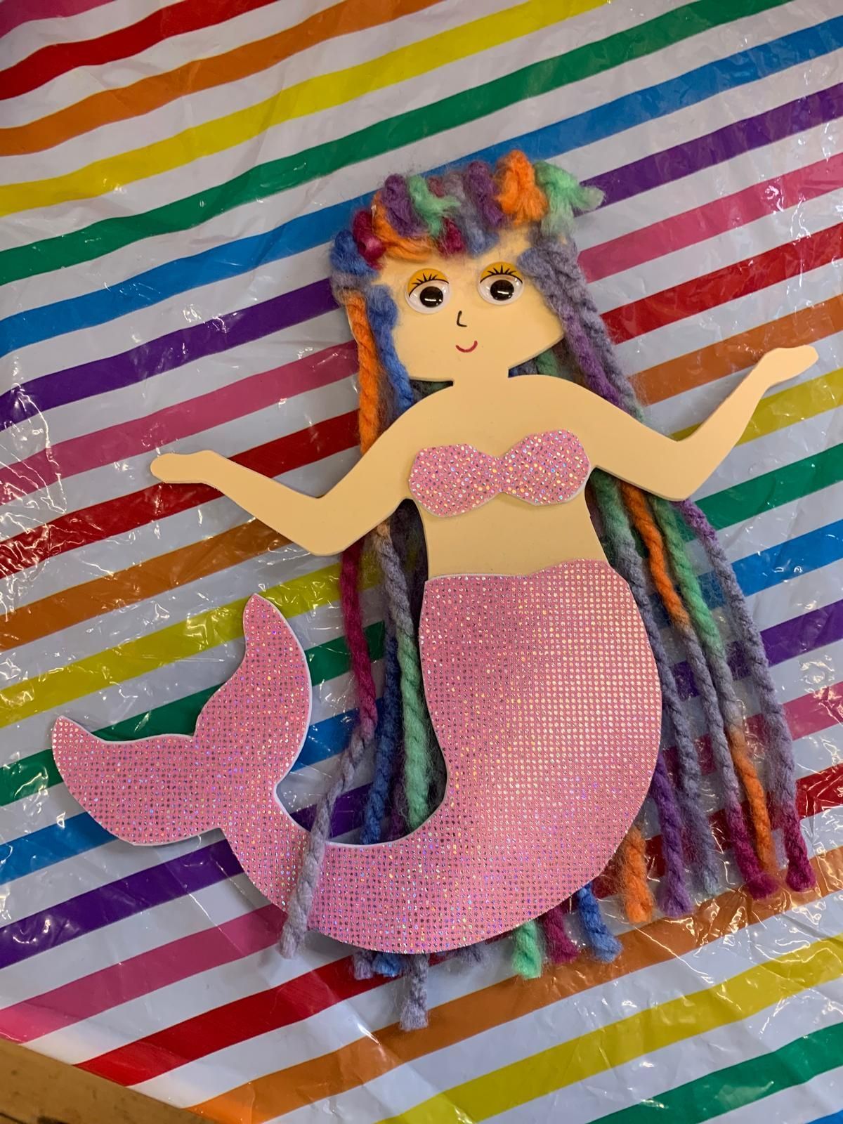 A paper mermaid with rainbow hair is sitting on a striped cloth for Dragonfly crafts party