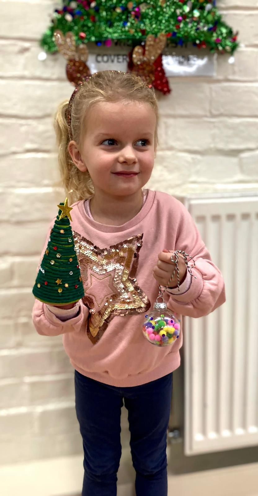 A little girl is holding a christmas tree in front of a christmas tree for Dragonfly crafts session