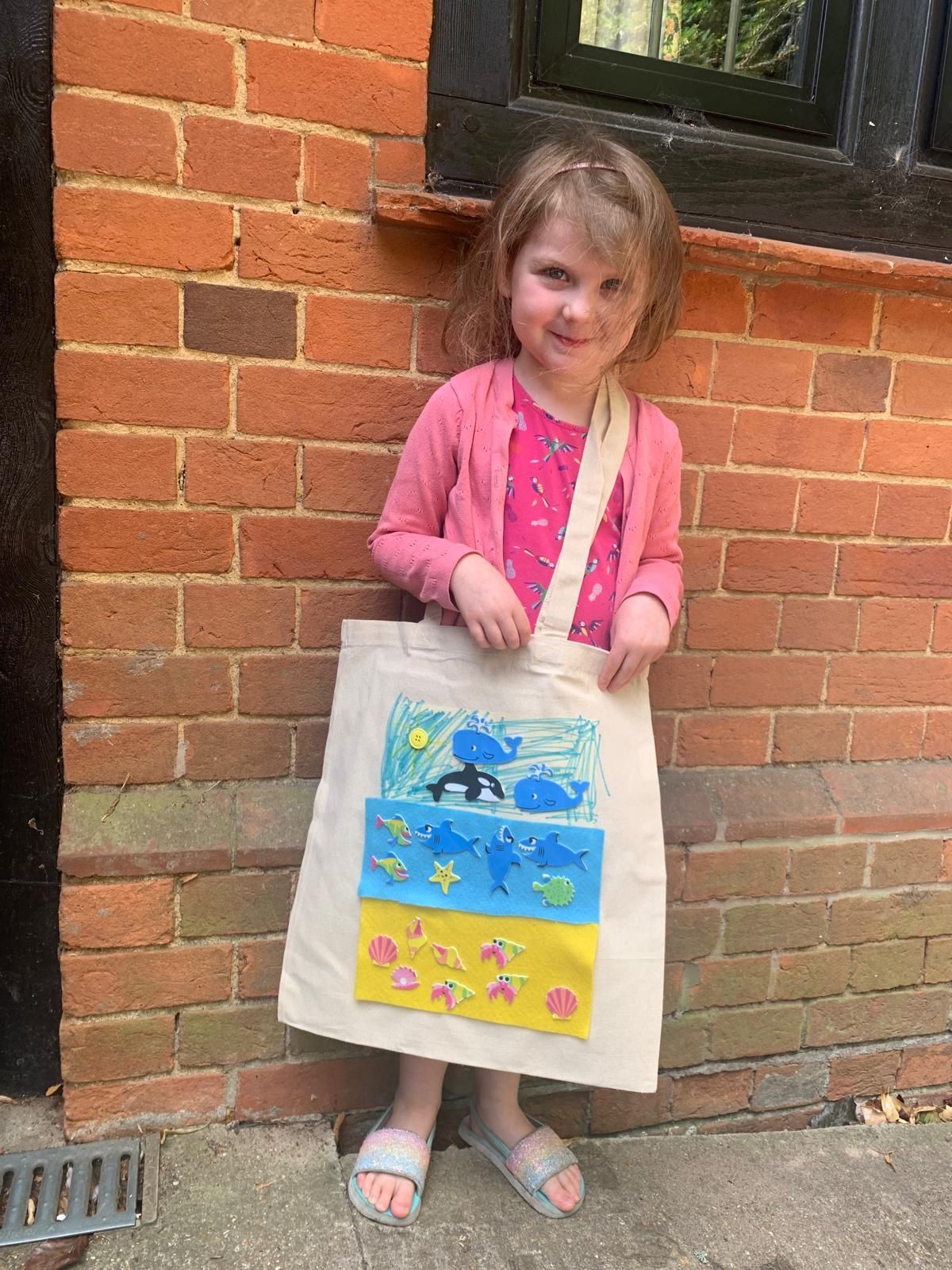 A little girl is standing in front of a brick wall holding a tote bag for Dragonfly crafts party