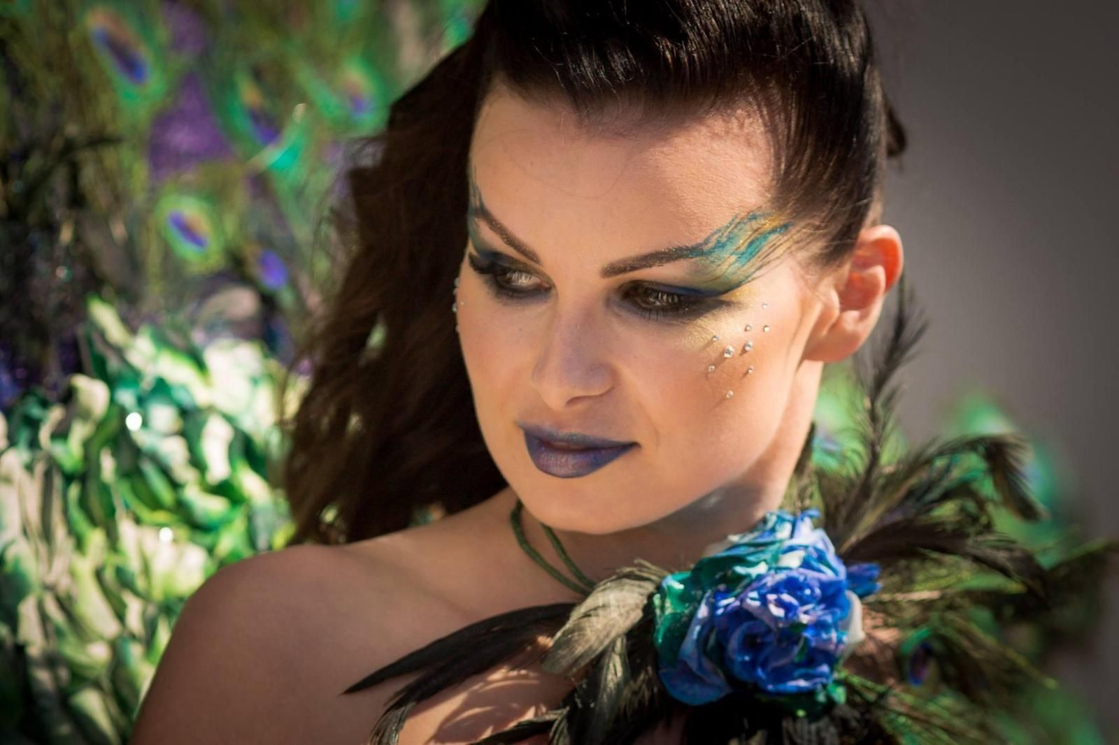 Una donna con piume di pavone dipinte sul viso tiene in mano una rosa blu.