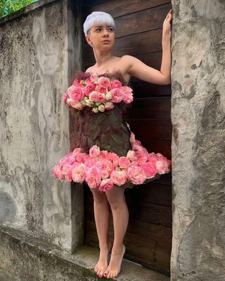 A woman in a dress made of flowers is standing next to a wall.