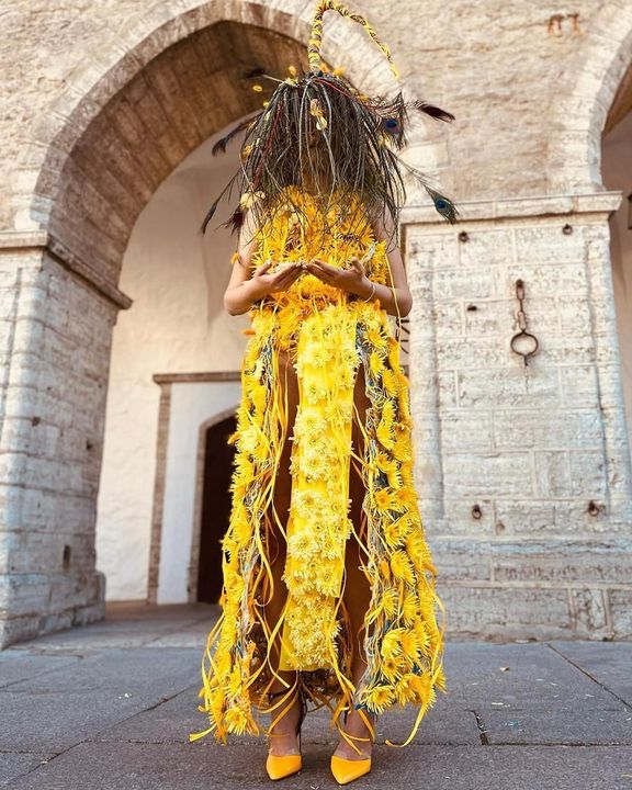 A woman with a yellow dress made of flowers is standing in front of a building