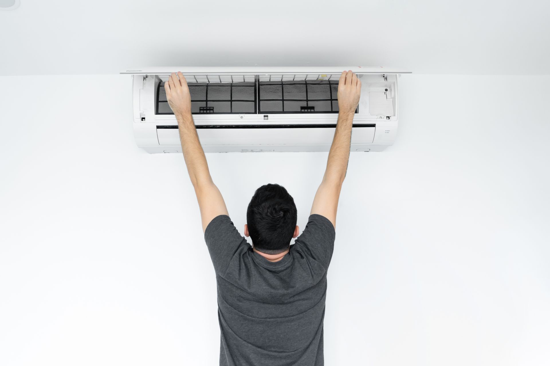 A man standing on his toes and reaching up to open the cover of a wall-mounted air conditioning unit