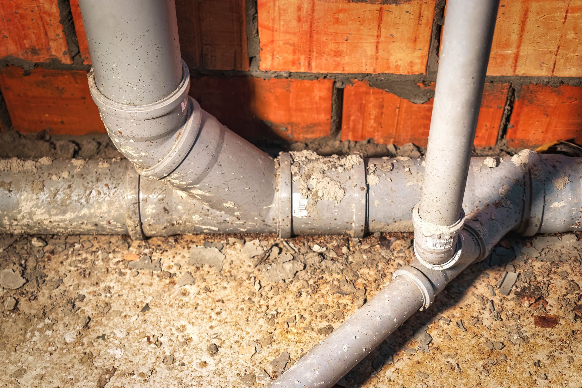 Gray PVC pipes running along a red brick wall on a concrete floor.