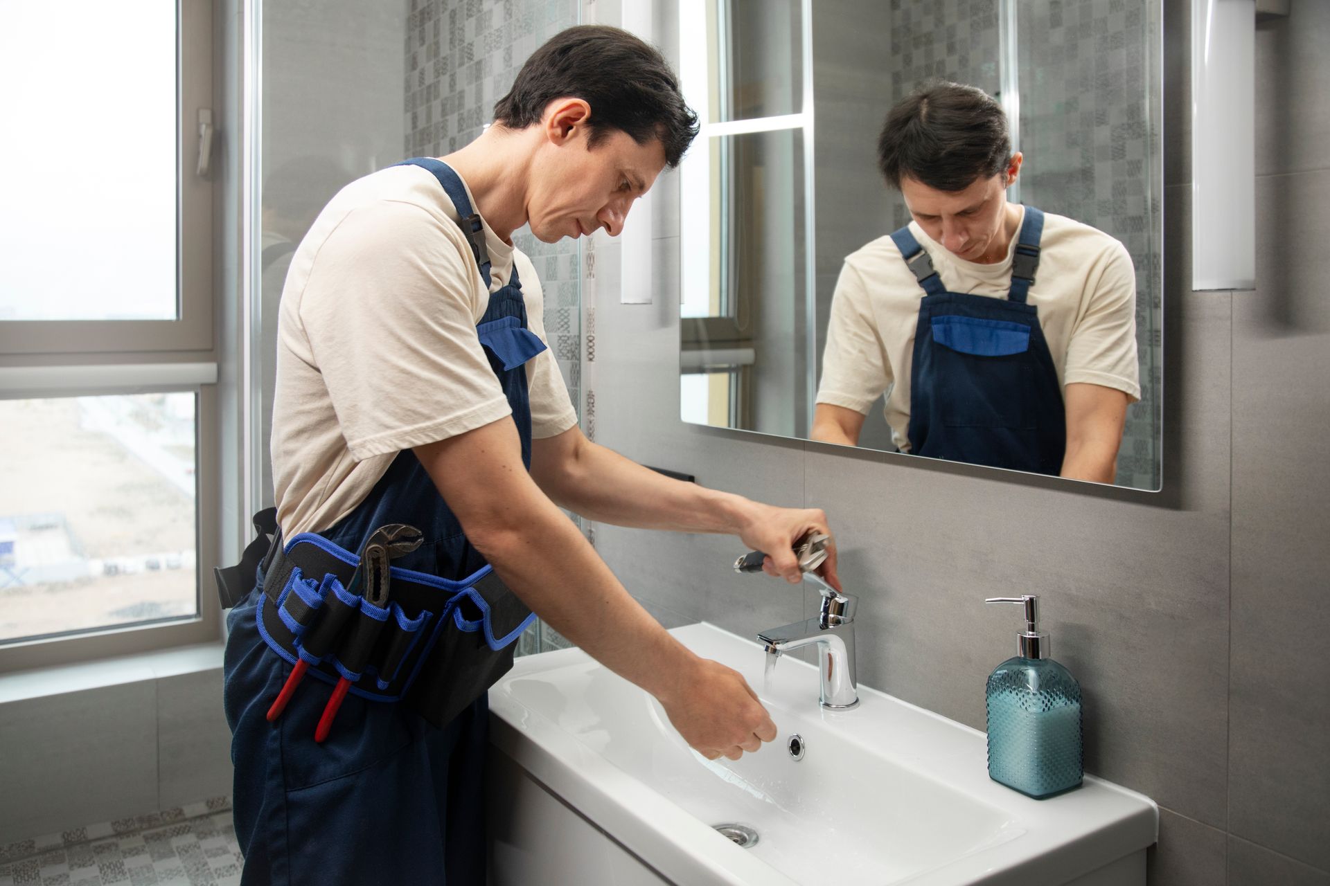 Person in blue overalls washing hands at a white bathroom sink with mirror reflection.