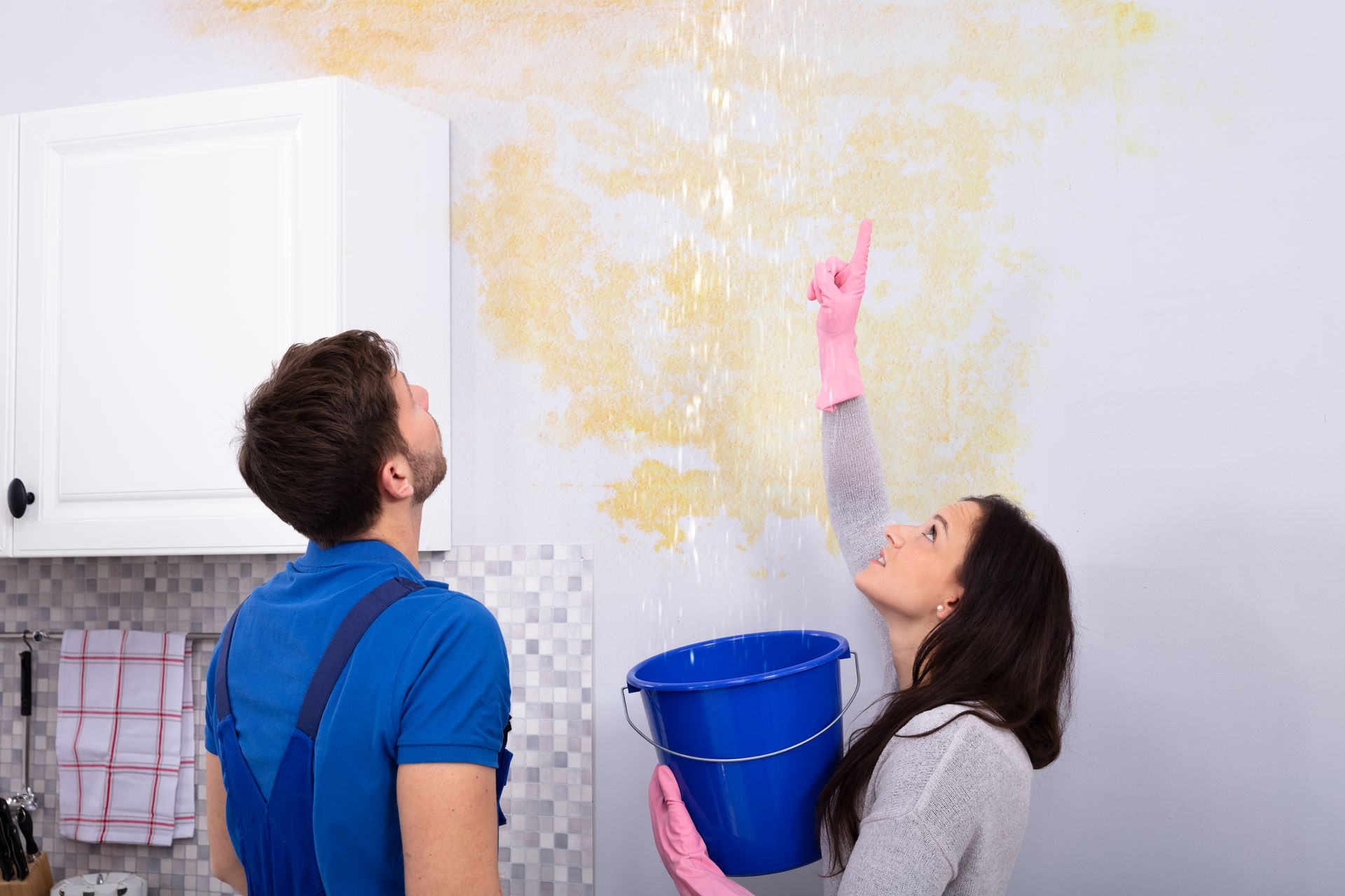 A man and a woman are looking at a leaking ceiling in a kitchen.