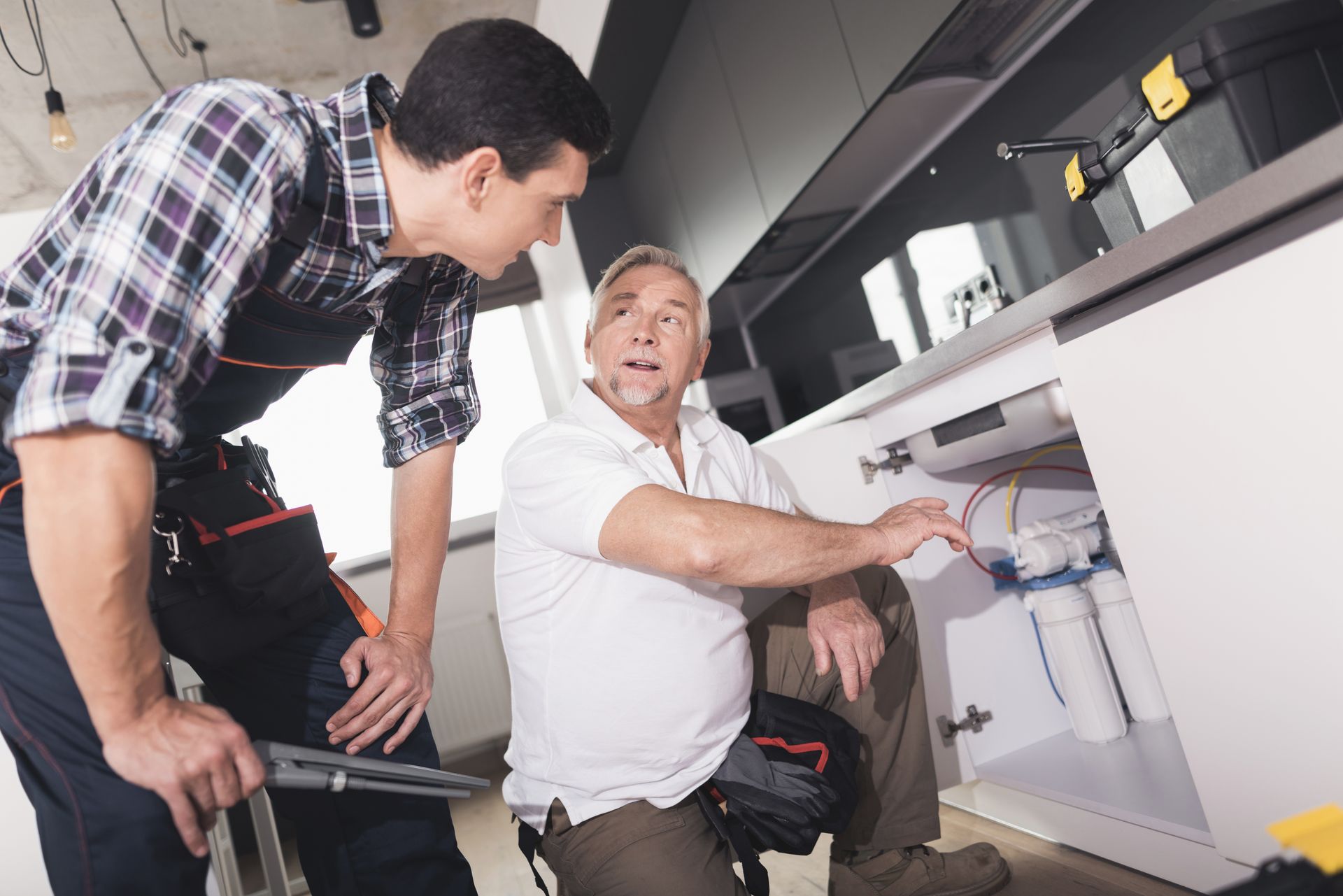 Two men are working on a sink in a kitchen.