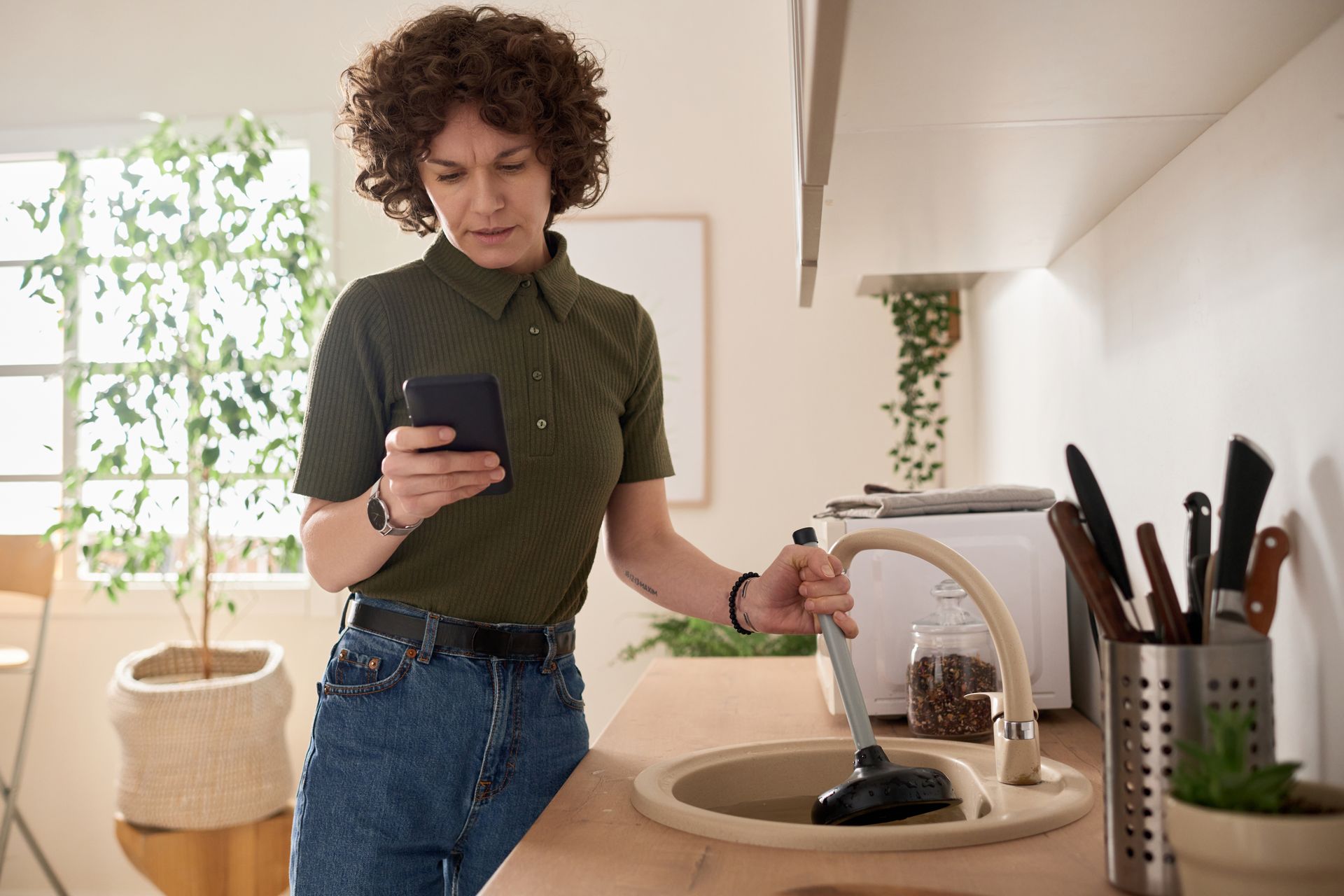 A woman is using a plunger to unblock a kitchen sink while looking at her phone.