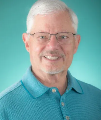 Smiling older man with glasses in a blue polo shirt, teal background.