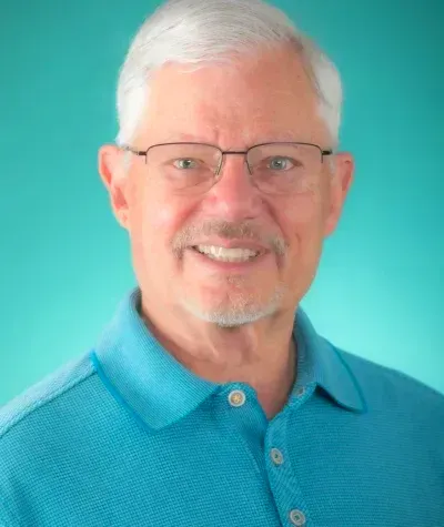 Smiling older man with glasses, blue shirt, teal background.