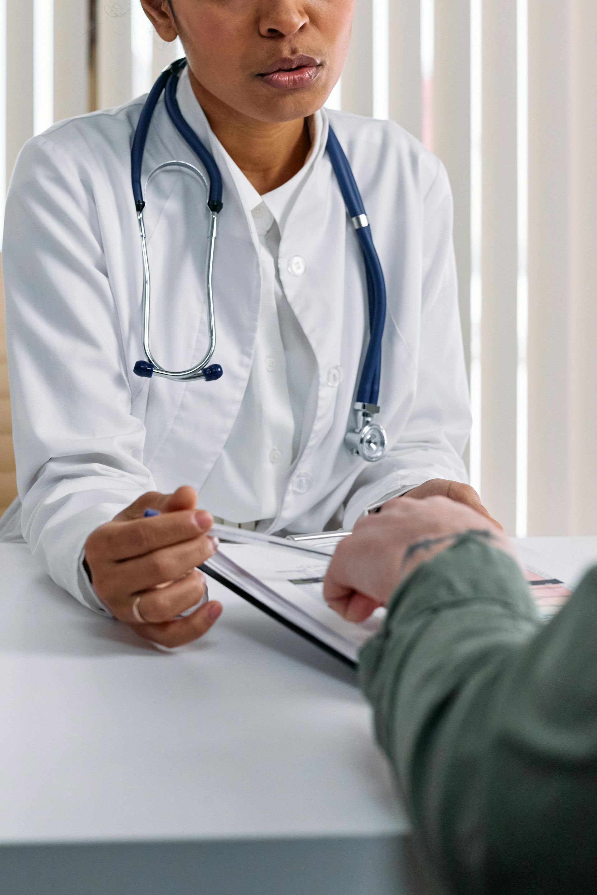 Doctor with stethoscope examines patient's wrist, sitting at a desk.