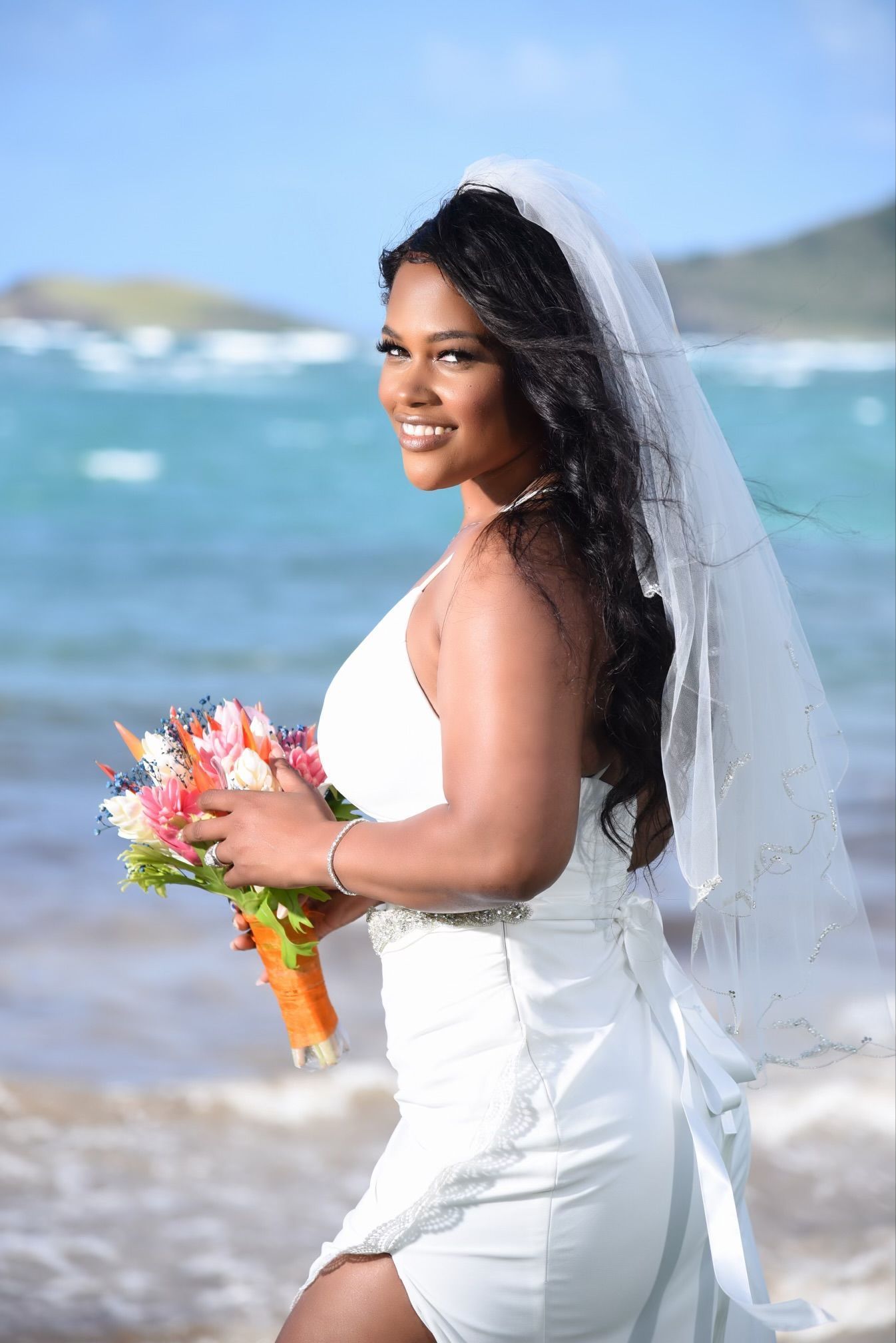 A bride in a white dress and veil is holding a bouquet of flowers on the beach.