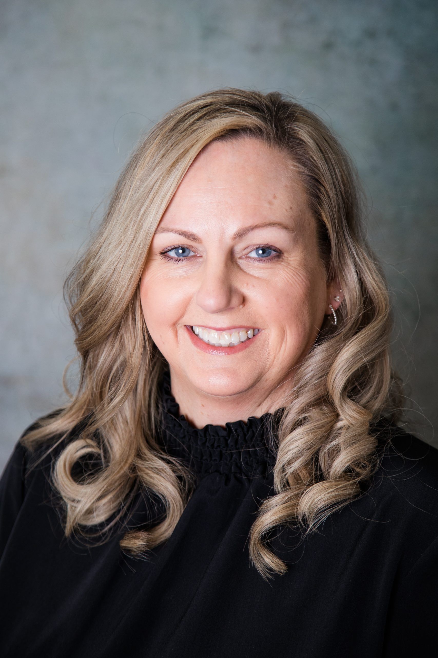 A woman with blonde hair and blue eyes is wearing a black sweater and smiling.