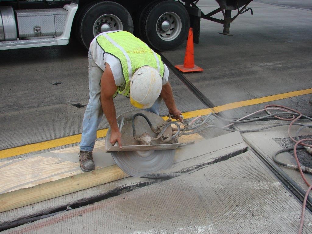 Worker using a circular saw to cut concrete on a road. They wear a safety vest and helmet.