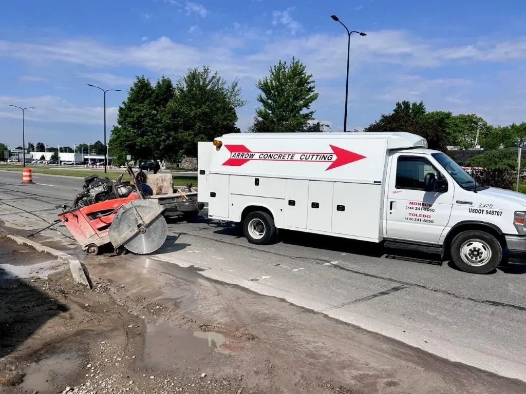 White work truck with red arrow logo, towing a road saw cutting asphalt.