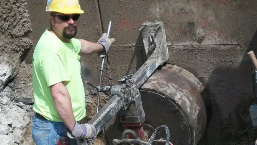 Construction worker in a yellow hard hat and sunglasses operates a machine. He's outside near a large metal container and dirt.