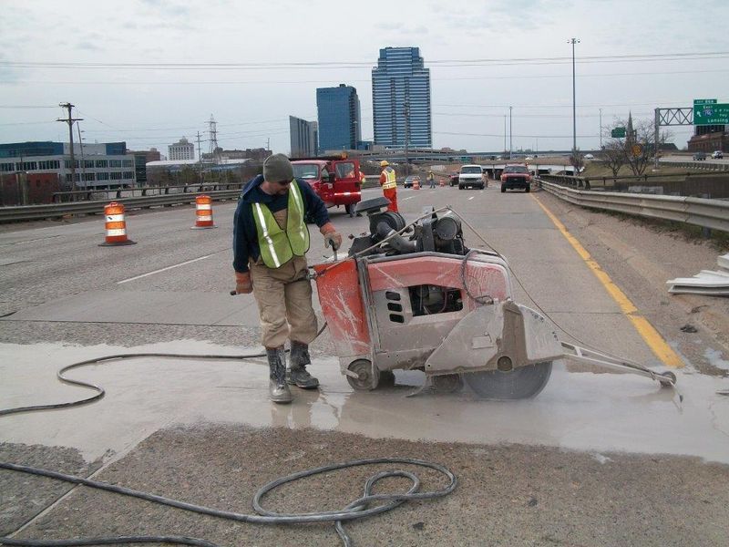 Man using a concrete saw to cut pavement on a highway, with city skyline in the background.